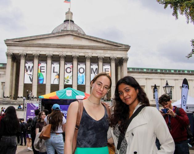 two students standing in front of crowd and tents with a large stone pillared building in the background that has the word "welcome" written on banners