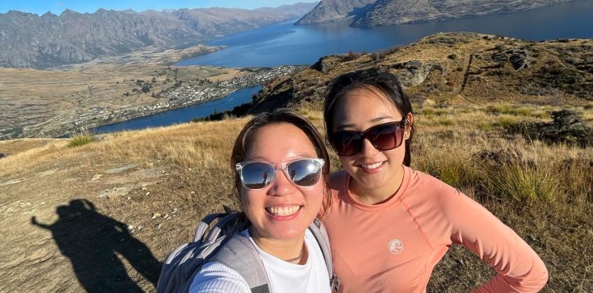 Two girls pose for a selfie at the top of the Queenstown Hill. The girl on the left wears a white shirt and jeans, and the girl on the right wears a pink shirt and black pants. The background shows a few mountains and a clear sky. 