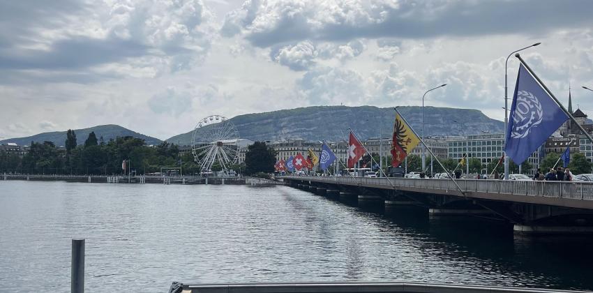 on the left side is a view of a large lake; on the right side is a bridge with various flags hanging over the side; the mountains of Switzerland are visible in the background