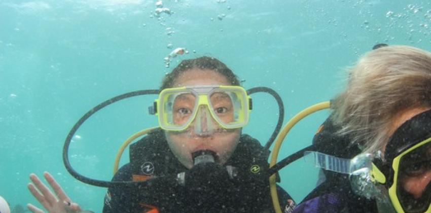 A girl waves at the camera as she scuba dives. She has goggles on her face and a mouthpiece helping her breathe. There is a fish in the bottom left corner.