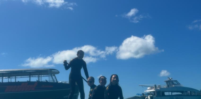 Three girls sit on a paddleboard while another girl stands up and paddles. Two boats are behind them, and the sky is clear with a few puffy clouds.