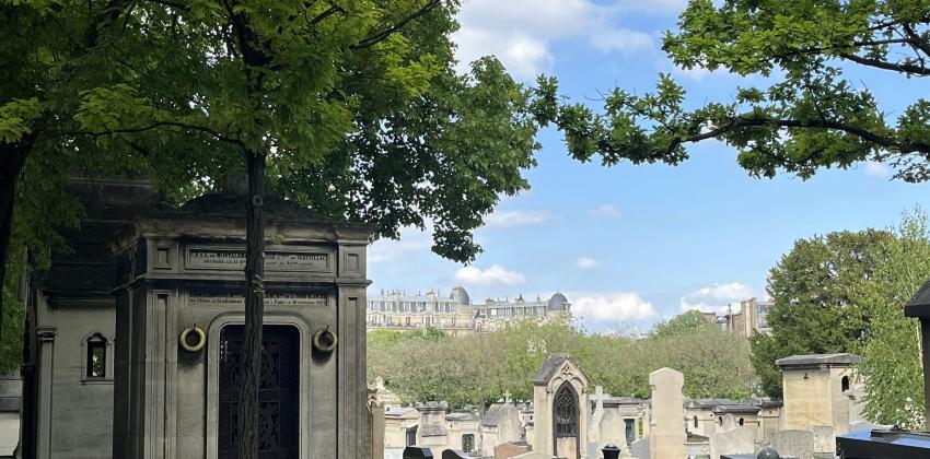 standing outside on the pavement in a cemetery; tombs and trees are visible, while there is a nice view of city buildings in the background