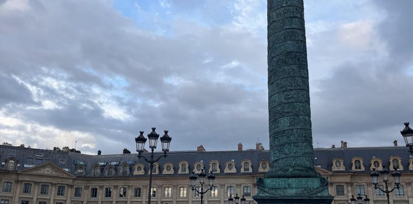 standing on pavement outside; in the background are large buildings with classic architecture, to the left in the foreground is a tall blueish column