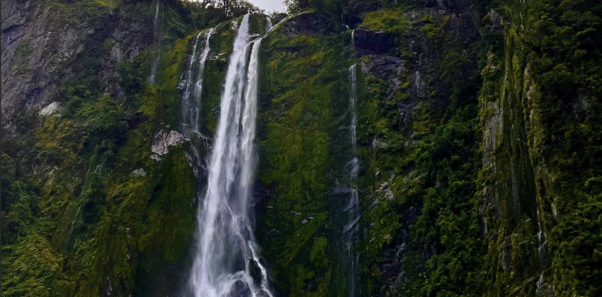 A large waterfall falls from a steep forested cliff. The sky is dark and cloudy.