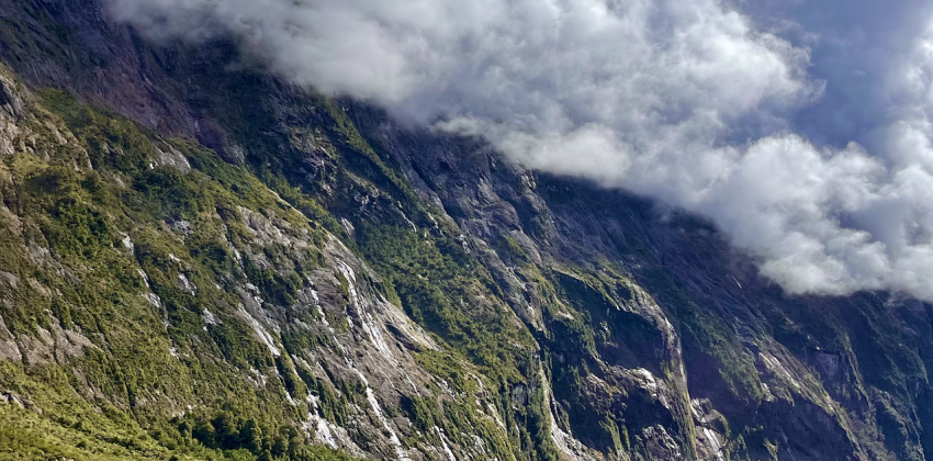 White clouds loom over the large mountain face of Milford Sound. There is a road that snakes through the landscape.