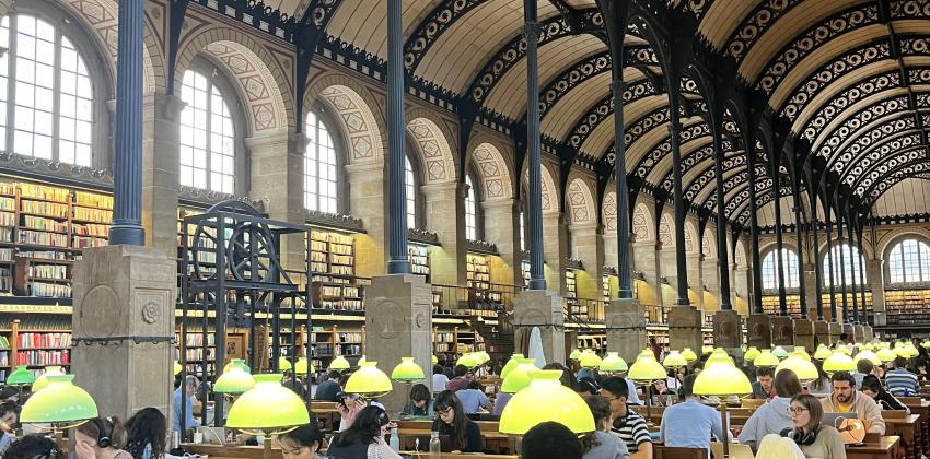 inside a library with tall ceilings and big arches; students are working at long wooden tables with lamps