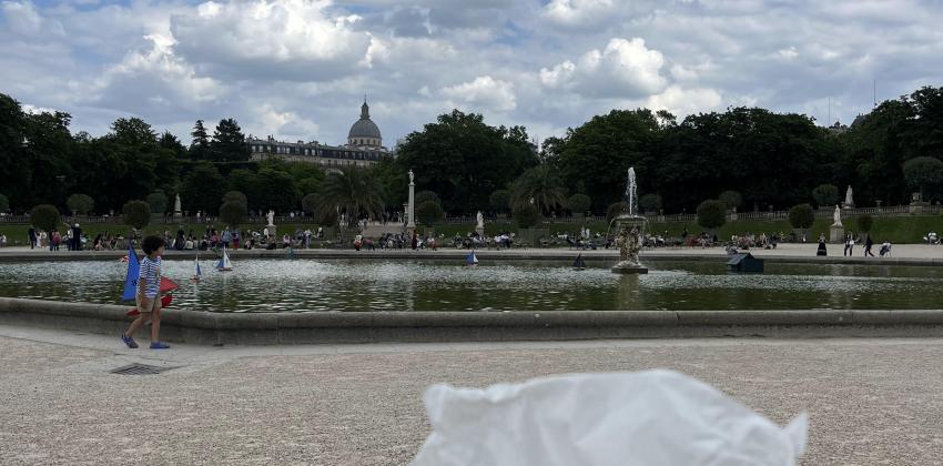 a hand is holding a cookie croissant while a vast garden is visible in the back; trees and a large fountain are visible in the background