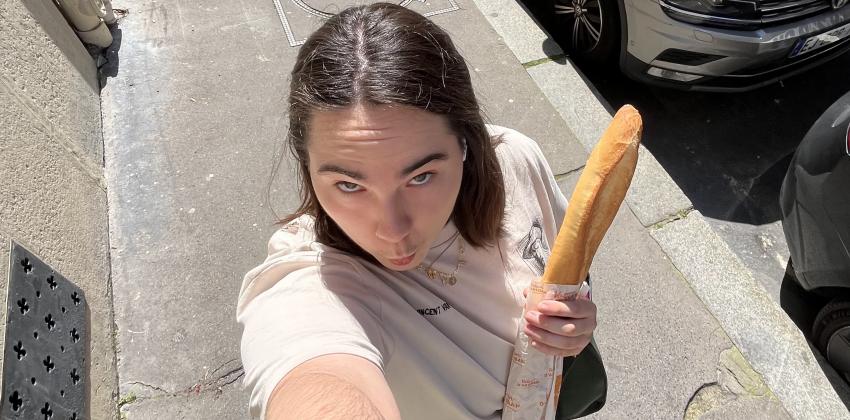 this is a 0.5 photo; there is a girl in the streets of Paris, holding a bag with a baguette and making a funny face