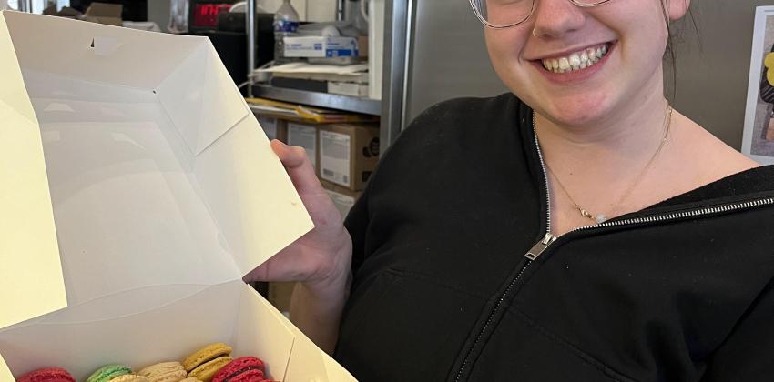 a photo inside a bakery kitchen; a girl is to the right and is holding a white box full of different types of macarons that she and her friends made for the first time