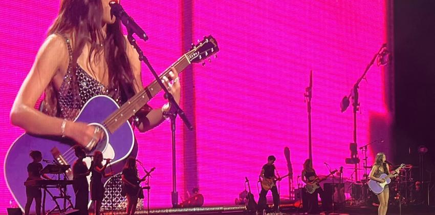 a photo of a stage in a large arena; Olivia Rodrigo is visible on screen and on the stage while playing the guitar