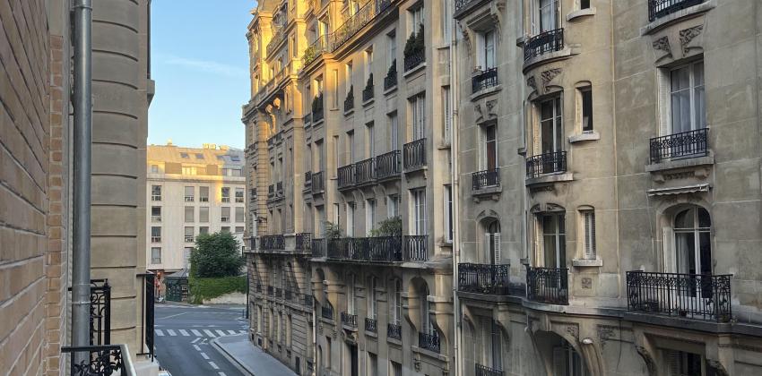 the photo is taken from the balcony of a residential building in Paris; it is early in the morning and the sun is rising, the sky is blue; there is a view down the street that included views of the buildings on the road