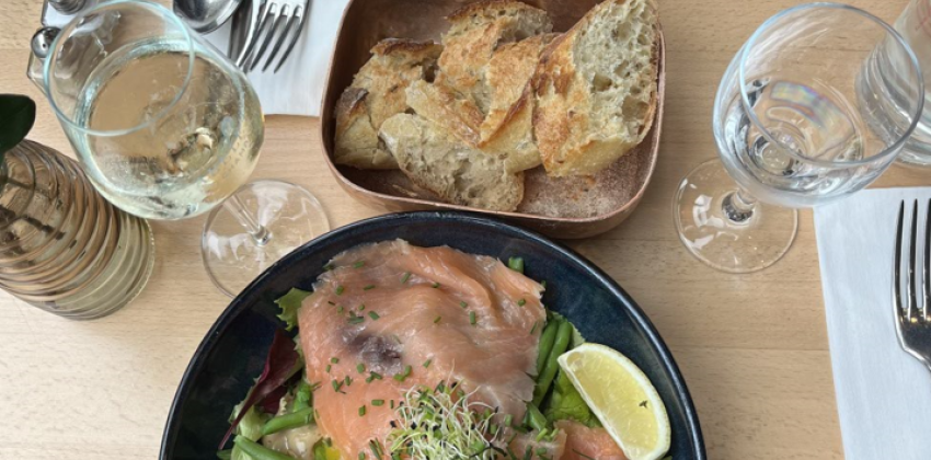 the photo is an overhead view of a table; there are two dishes, a basket of bread in the middle, two wine glasses and two sets of silverware and napkins; the table is a light wood color and the setting is well lit