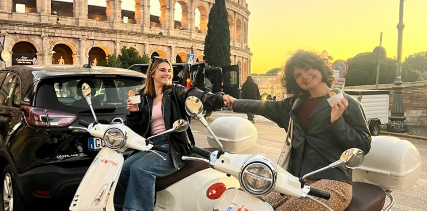 Atop white vespa scooters, Haley and Brooke sit and smile at the camera, holding hands and gelato cups. Behind them is the setting sun and the lit-up Colosseum.