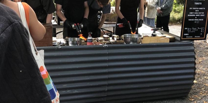 A picture of a farmer's market booth, with two workers making rib eye steak sandwiches.