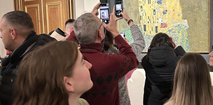 An average height white person with shoulder length brown way hair in a green t-shirt with a burnt orange crossbody bag looks at Klimt's Der Kuss. There are many other people in the background looking at the masterpiece and taking pictures of it.