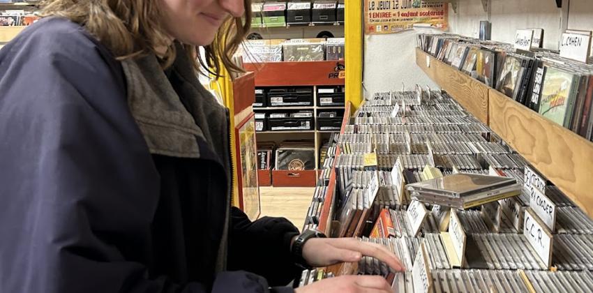 A white woman with shoulder length brown wavy hair in side profile wearing an oversized navy jacket looks at CDs in a record store