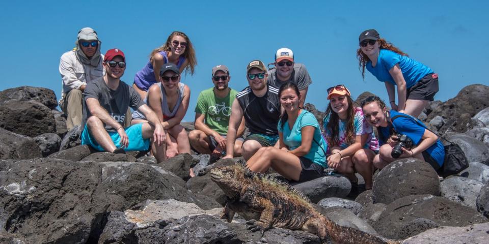 a group of students posing for a photo atop volcanic rock with a marine iguana